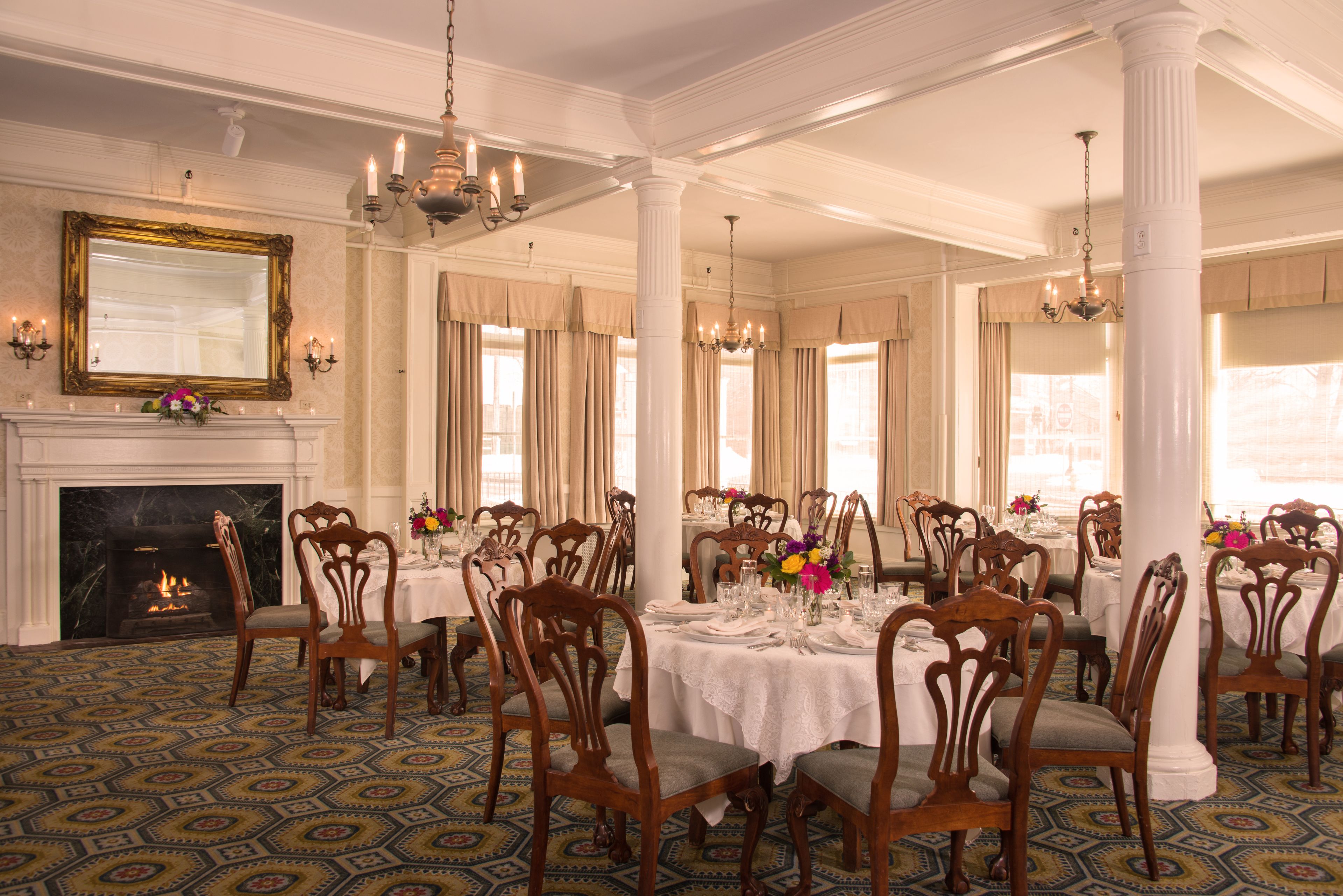 A formal dining room with multiple tables set for a meal, wooden chairs, a fireplace, and large windows with curtains.