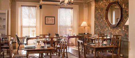 A dining area with multiple wooden tables and chairs, large windows with blinds, a chandelier, and a floral wallpaper.