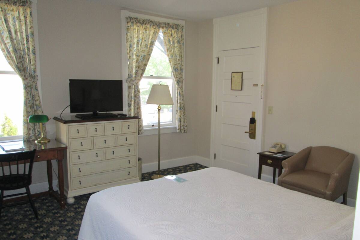 A hotel room with a bed, a desk with a computer, a chair, a lamp, and a television.