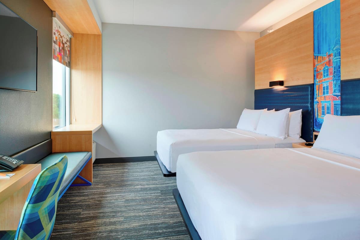 A hotel room with two beds, a desk with a chair, a flat-screen TV, and a large window.
