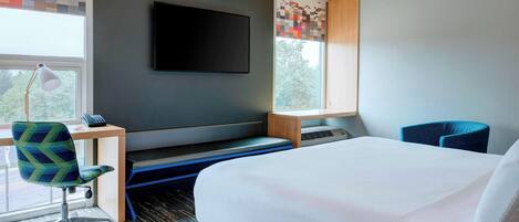 A modern hotel room with a large bed, a desk with a chair, a flat-screen TV mounted on the wall, and a window with a view of trees.