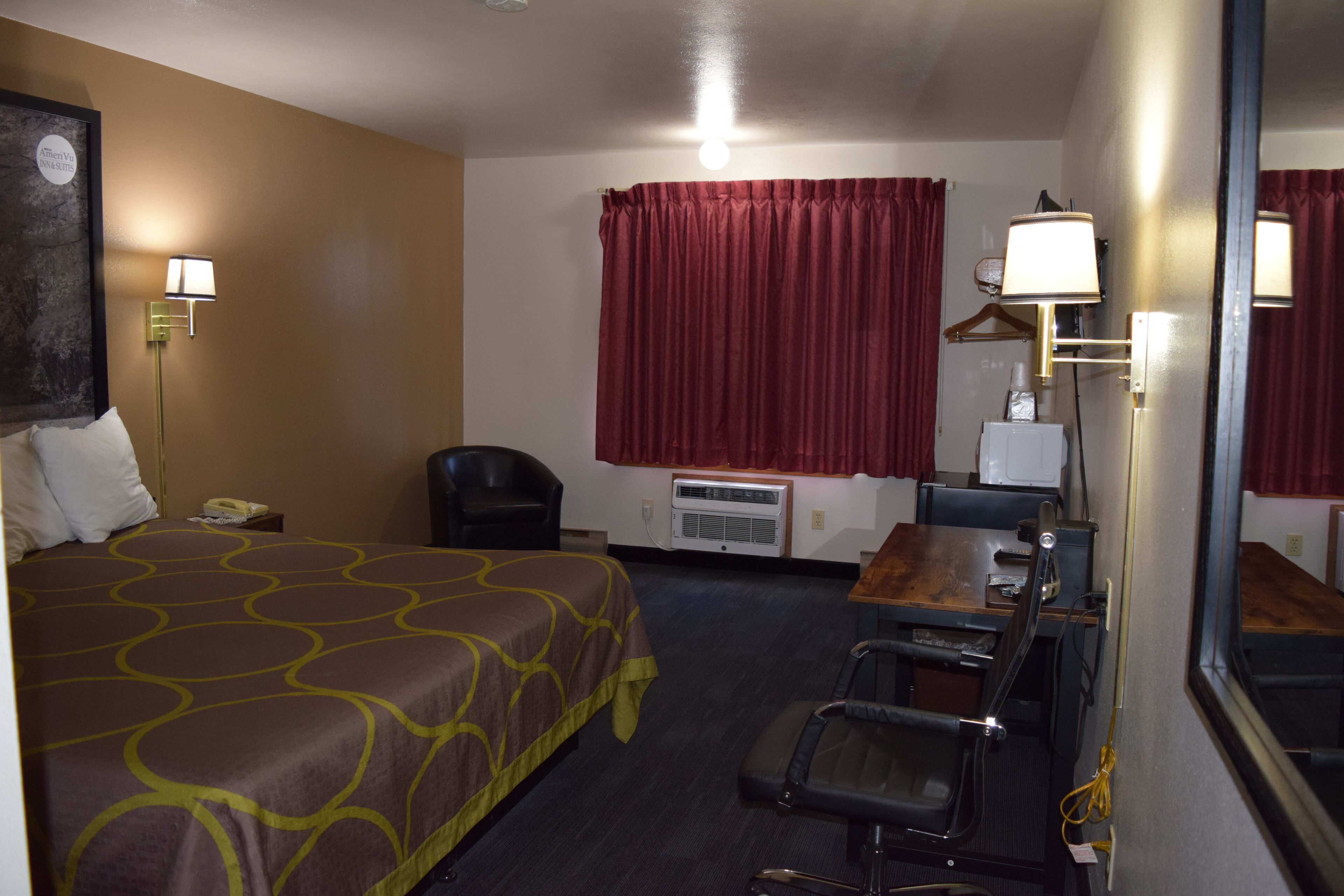 Habitación de hotel con una cama, un escritorio con silla, un espejo, una cortina roja y una lámpara fijada a la pared.