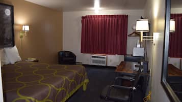 Habitación de hotel con una cama, un escritorio con silla, un espejo, una cortina roja y una lámpara fijada a la pared.