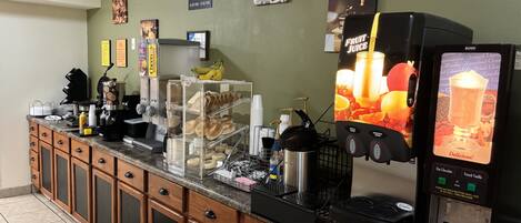 Una barra de desayuno con una máquina de café, un dispensador de zumo y una selección de bollería.