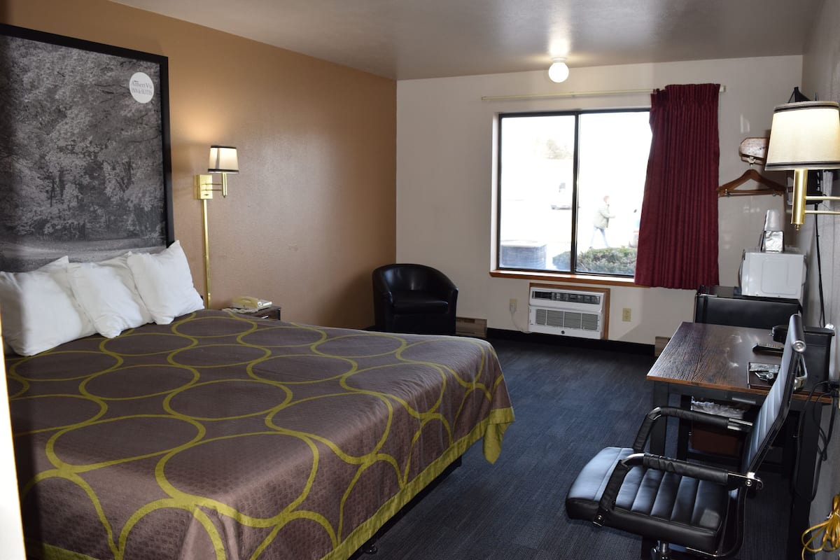 A hotel room with a large bed, a desk with a chair, a window with a view, and a wall-mounted lamp.