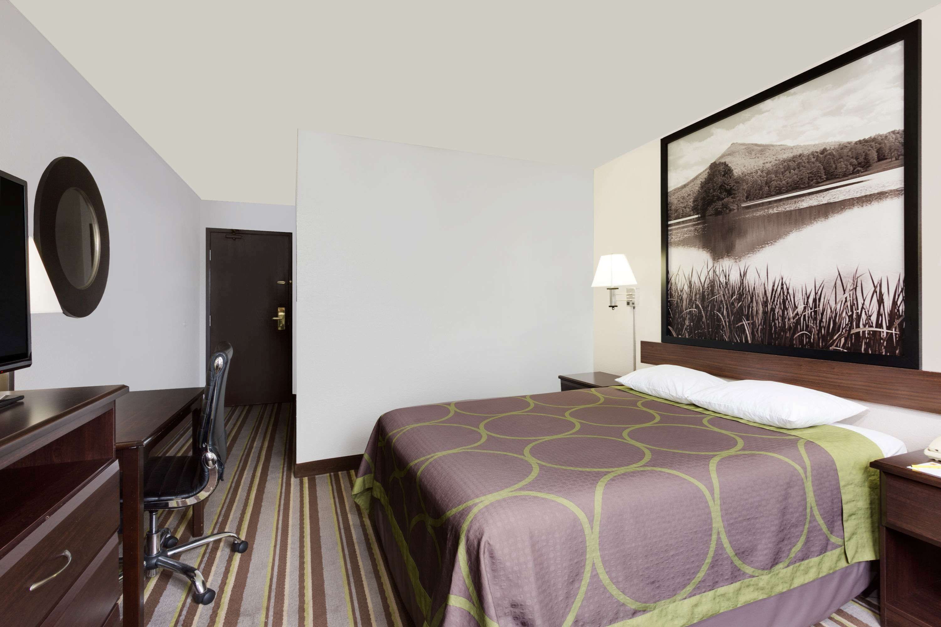 Een hotelkamer met een bed, een bureau, een stoel, een televisie, een lamp en een schilderij van een meer.