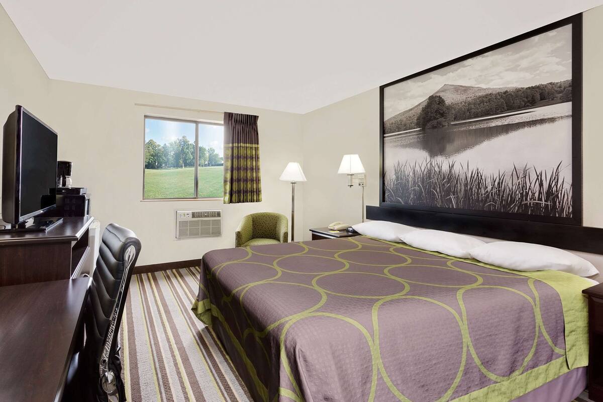 A hotel room with a bed, a desk with a TV, a chair, a window with curtains, and a painting of a lake.