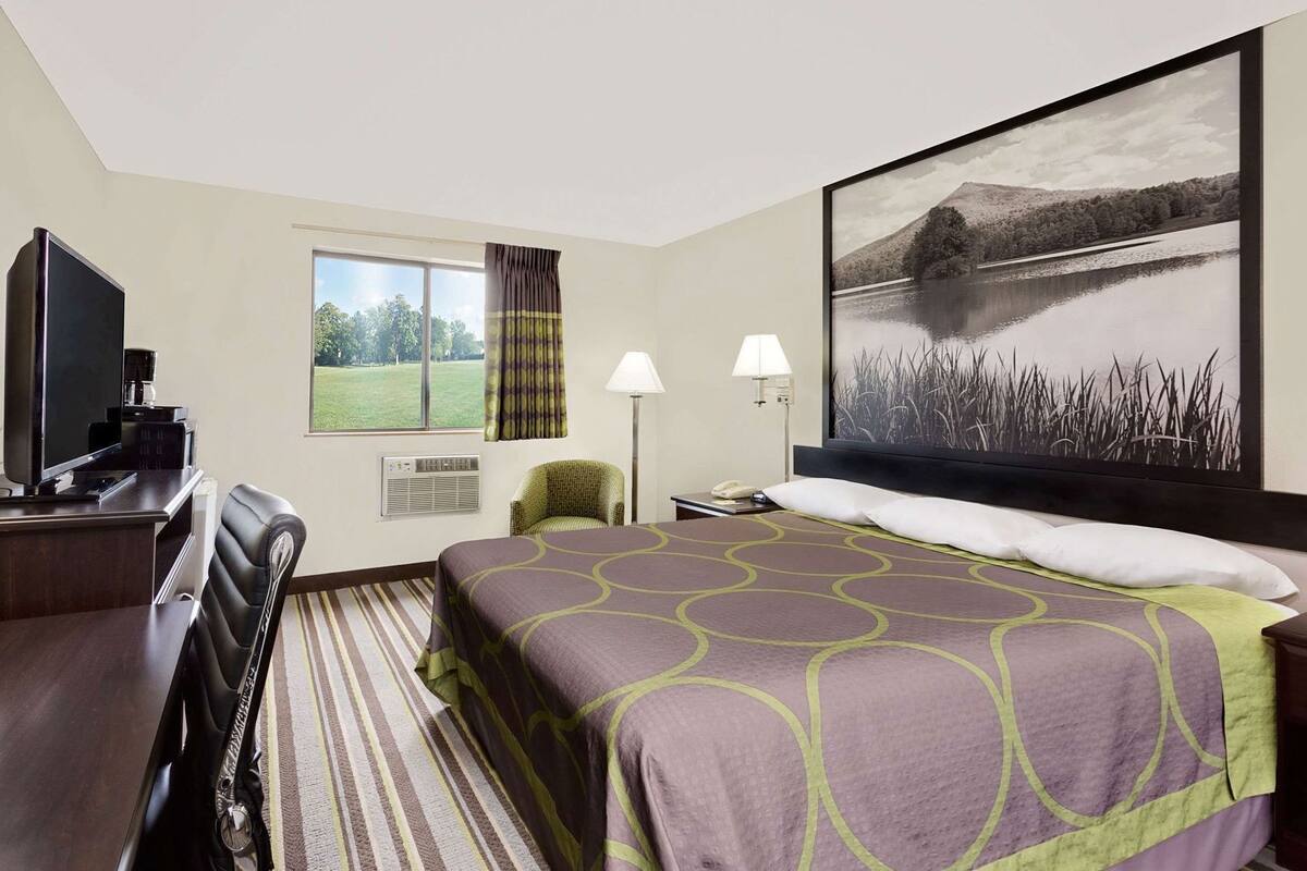 A hotel room with a bed, a desk, a TV, a chair, a lamp, and a large window with curtains.