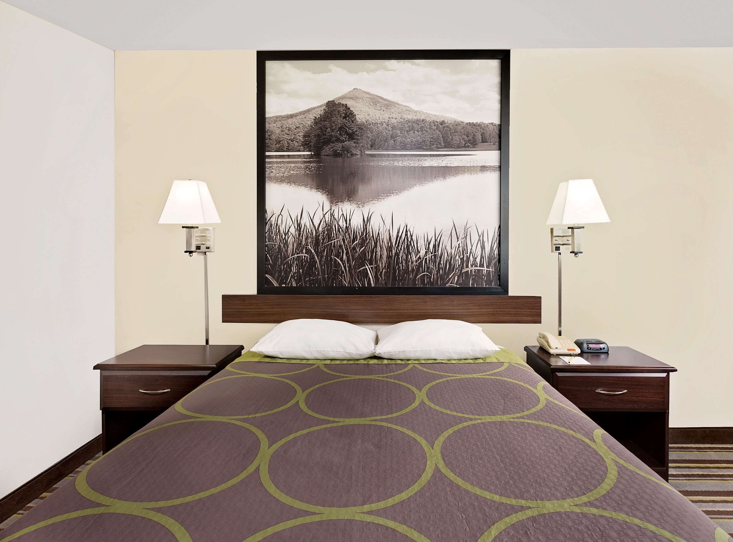 A hotel room with a bed, two bedside tables, a nightstand, a lamp, and a large framed picture of a lake and mountain.
