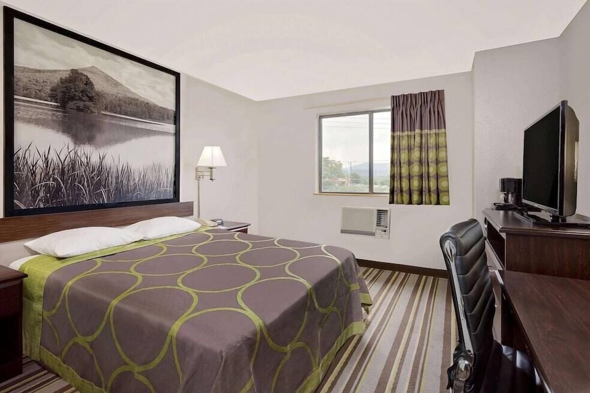 A hotel room with a bed, a desk, a television, a window with curtains, a painting, and an air conditioning unit.