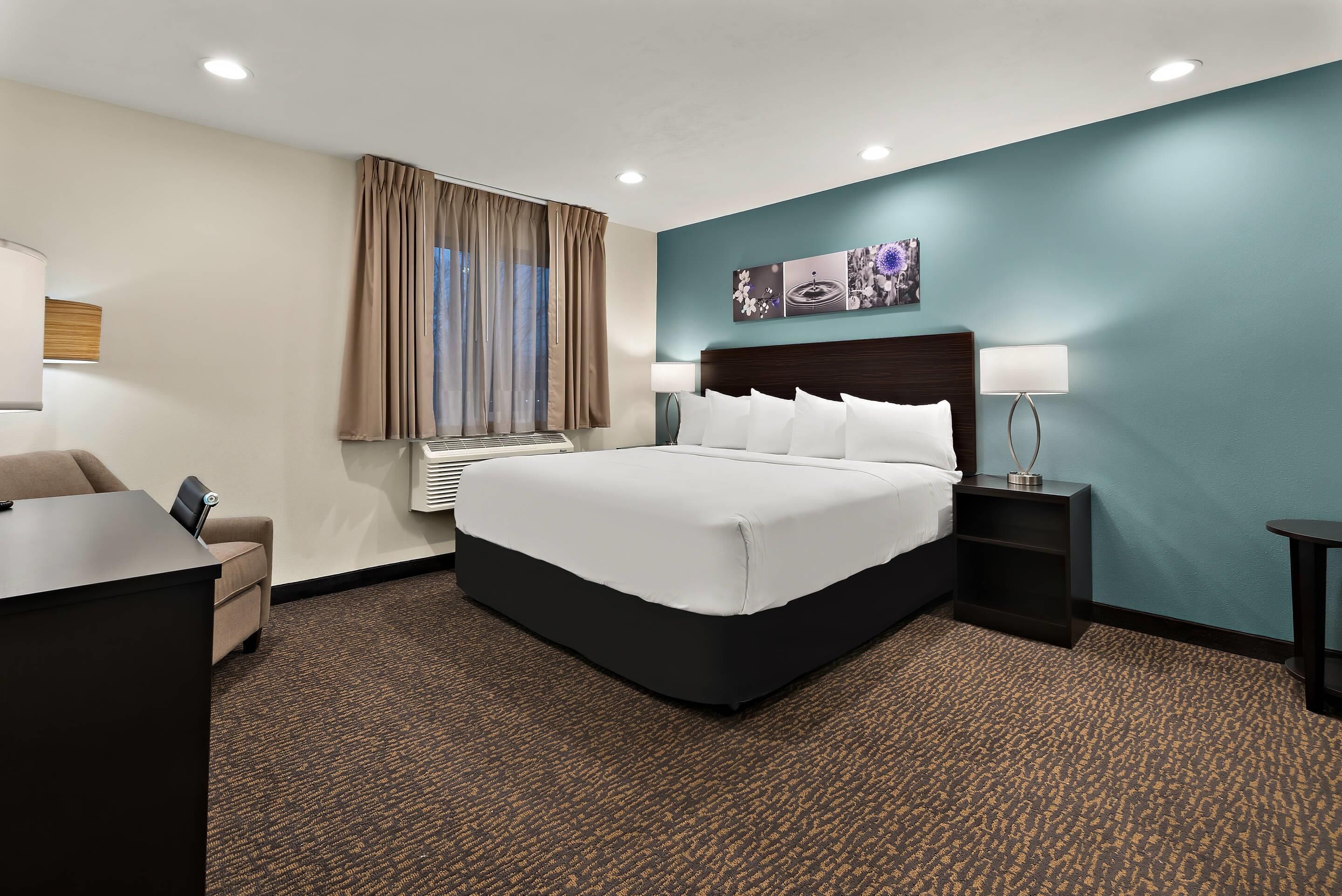 A hotel room with a large bed, a desk, a chair, a nightstand, a lamp, and a wall-mounted air conditioner.