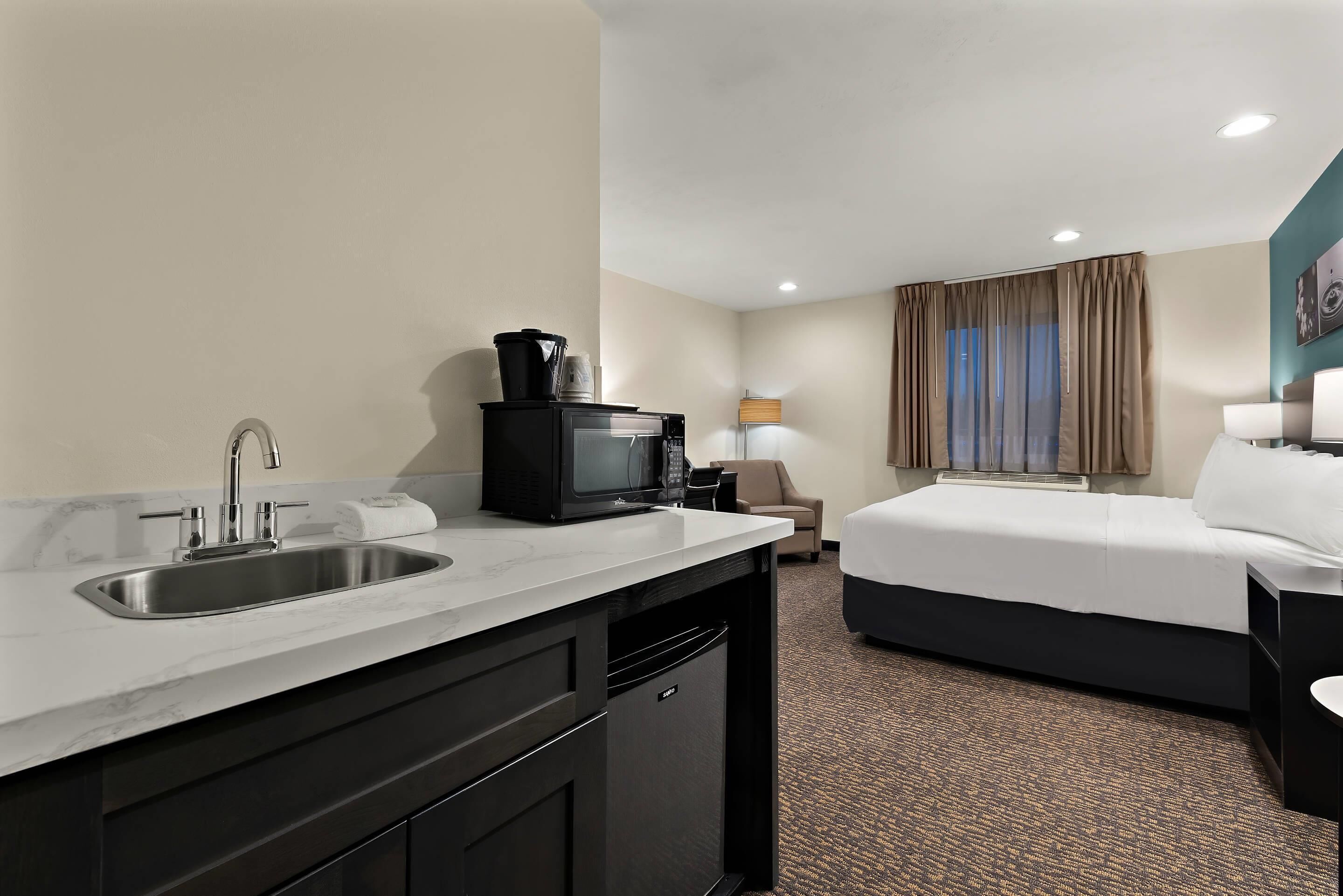 A hotel room with a kitchenette, a bed, a desk, and a lamp.