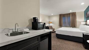 A hotel room with a kitchenette, a bed, a desk, and a lamp.