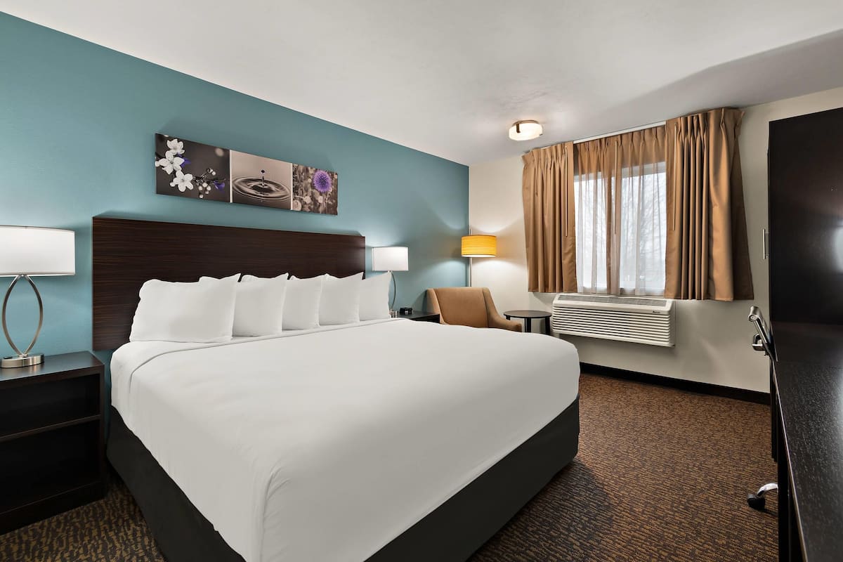 A hotel room with a large bed, a nightstand with a lamp, a chair, a desk with a lamp, a window with curtains, and artwork on the wall.