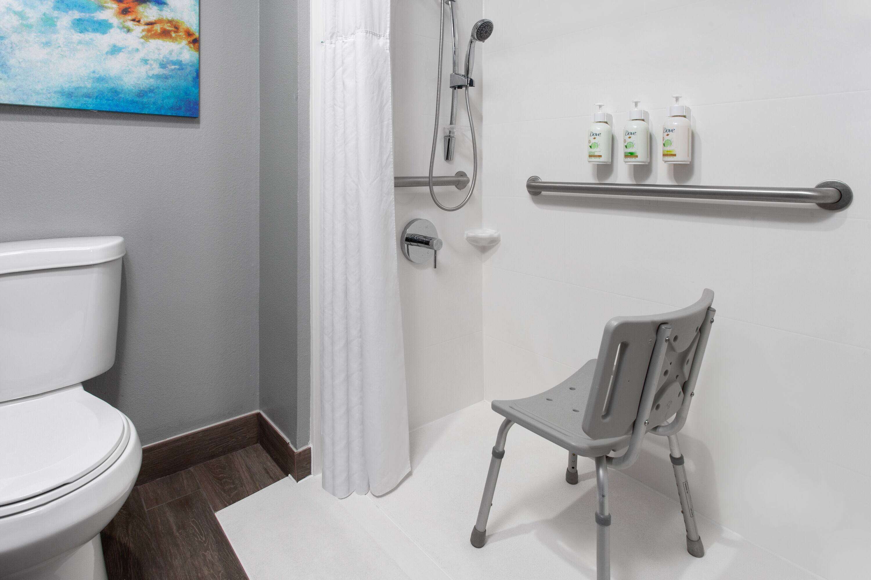 A bathroom with a white toilet, a shower with a handheld showerhead, a grab bar, and three bottles of Dove soap on a shelf.