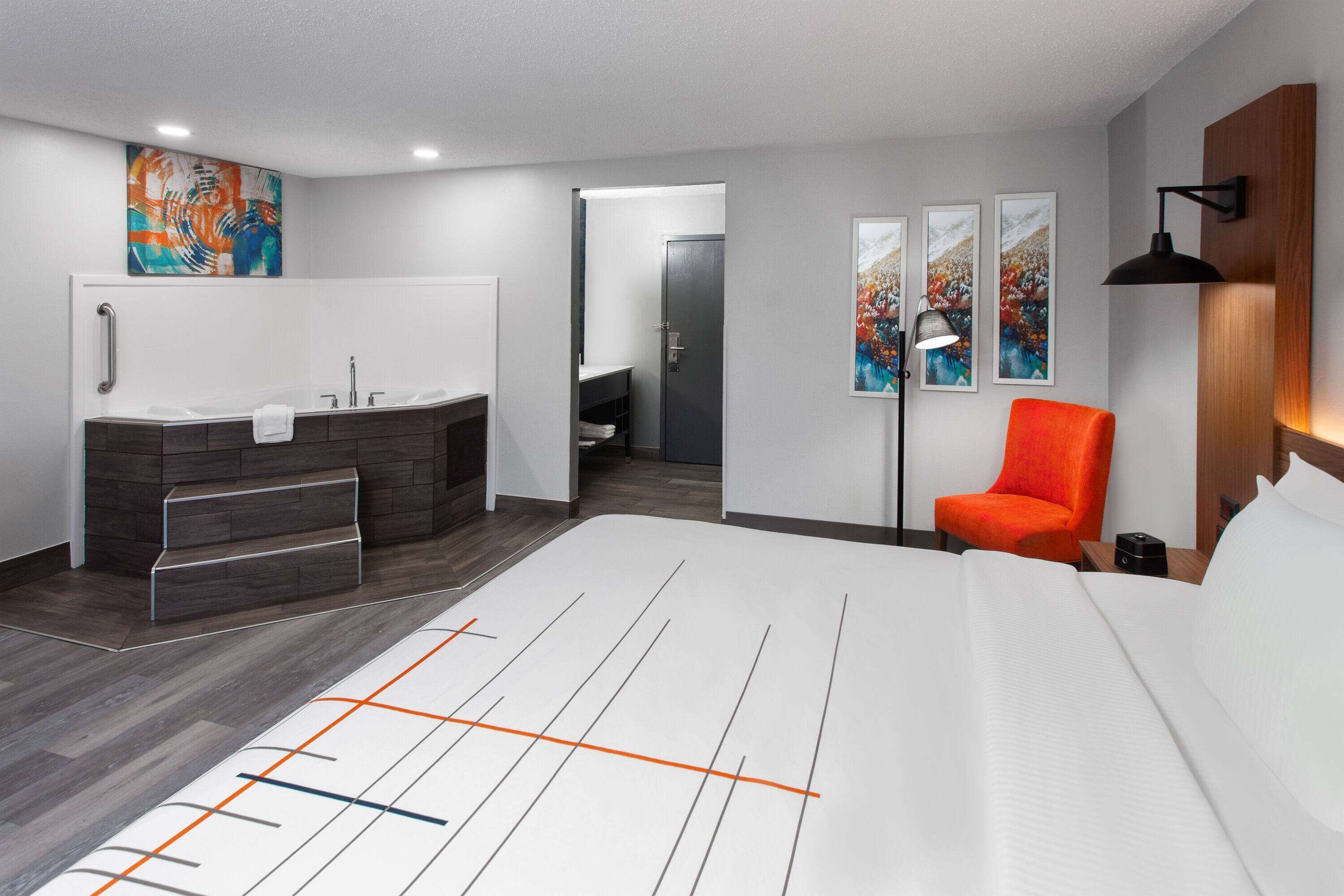 A modern hotel room with a large bed, a bathroom with a bathtub, a chair, and artwork on the walls.