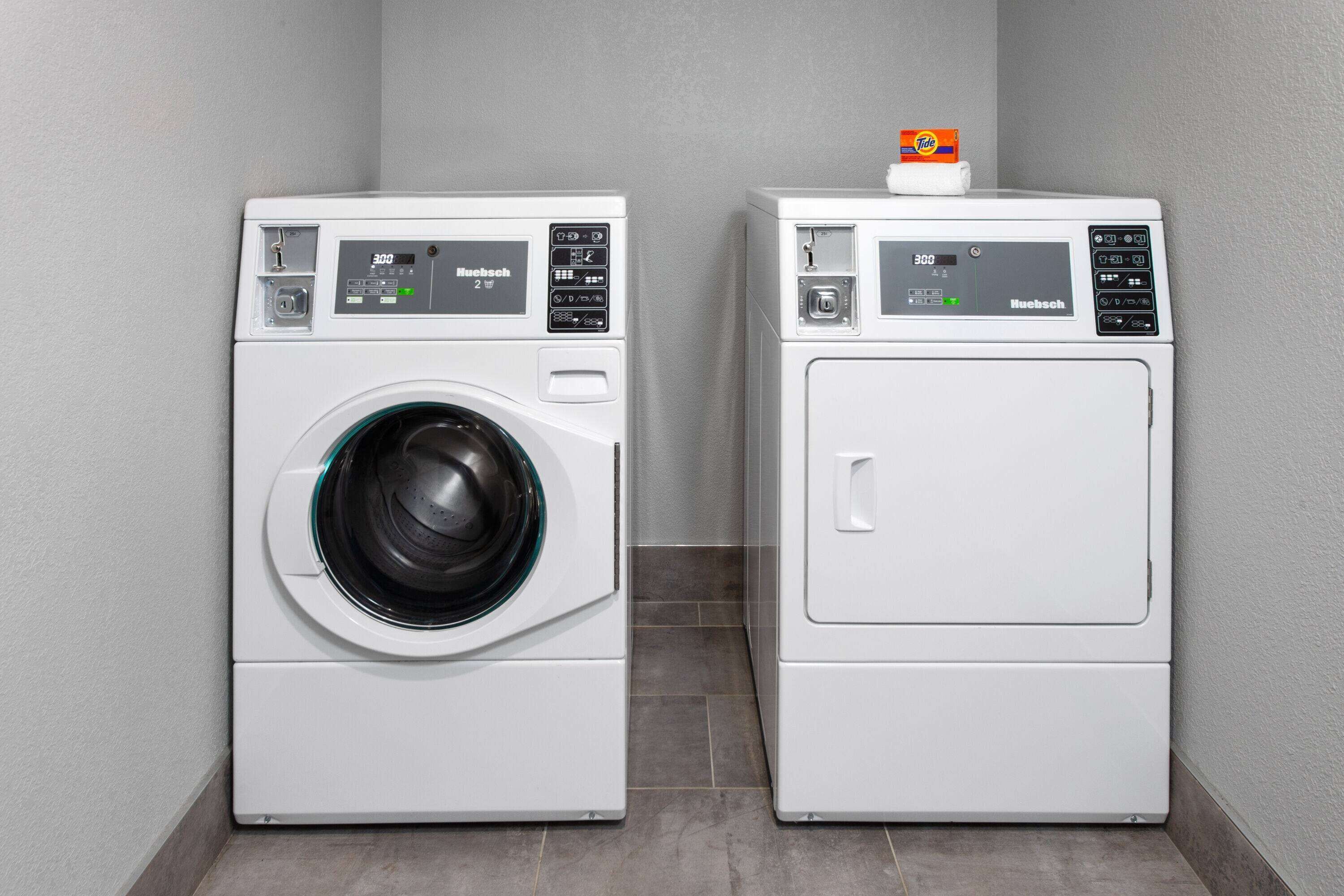 Two white, modern front-loading washing machines with digital displays, placed in a corner with a grey tiled floor and walls.