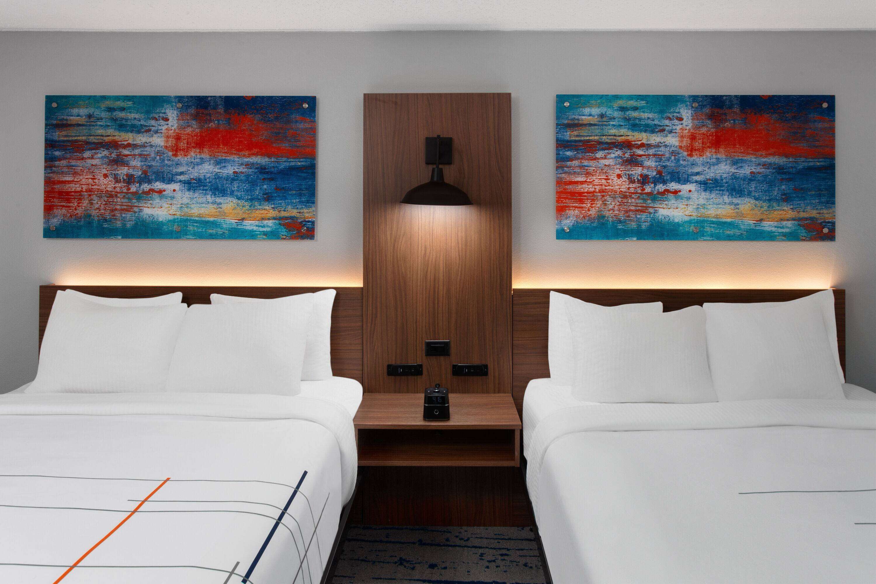 A hotel room with two beds, a wooden headboard, a nightstand, a lamp, and abstract paintings on the walls.