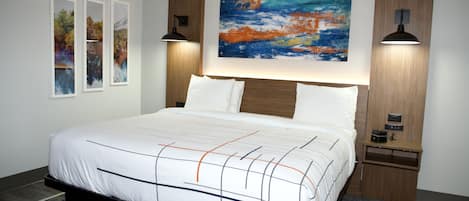 A modern hotel room with a large bed, a nightstand with a lamp, a wall-mounted artwork, and framed pictures on the wall.