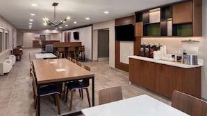 Un comedor moderno de hotel con una larga mesa de madera, varias sillas, una estación de café con diversas bebidas y un televisor montado en la pared.