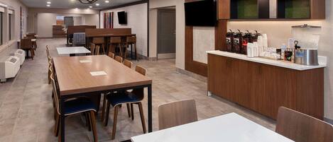 A modern hotel dining area with a long wooden table, several chairs, a coffee station with various beverages, and a TV mounted on the wall.
