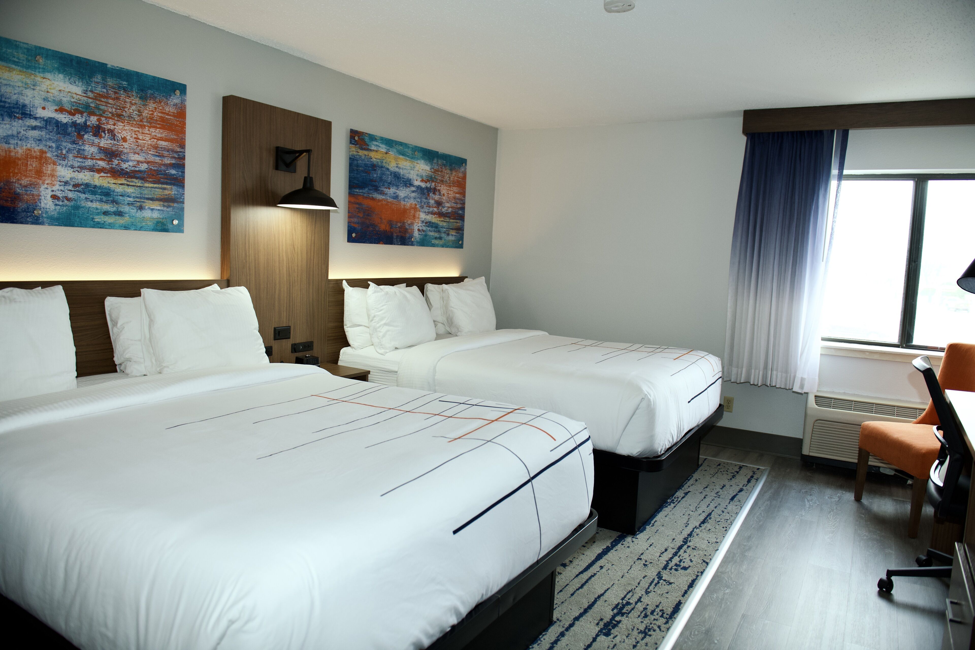 Ein Hotelzimmer mit zwei Betten, einem Schreibtisch und einem Stuhl. An der Wand befinden sich zwei Gemälde und ein Fenster mit Vorhängen.