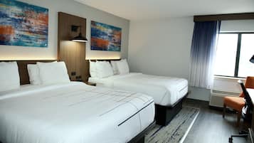 A hotel room with two beds, a desk, and a chair. There are two paintings on the wall and a window with curtains.