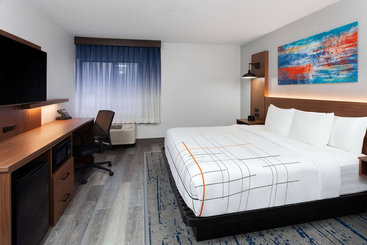 A hotel room with a bed, desk, chair, TV, and artwork on the wall.