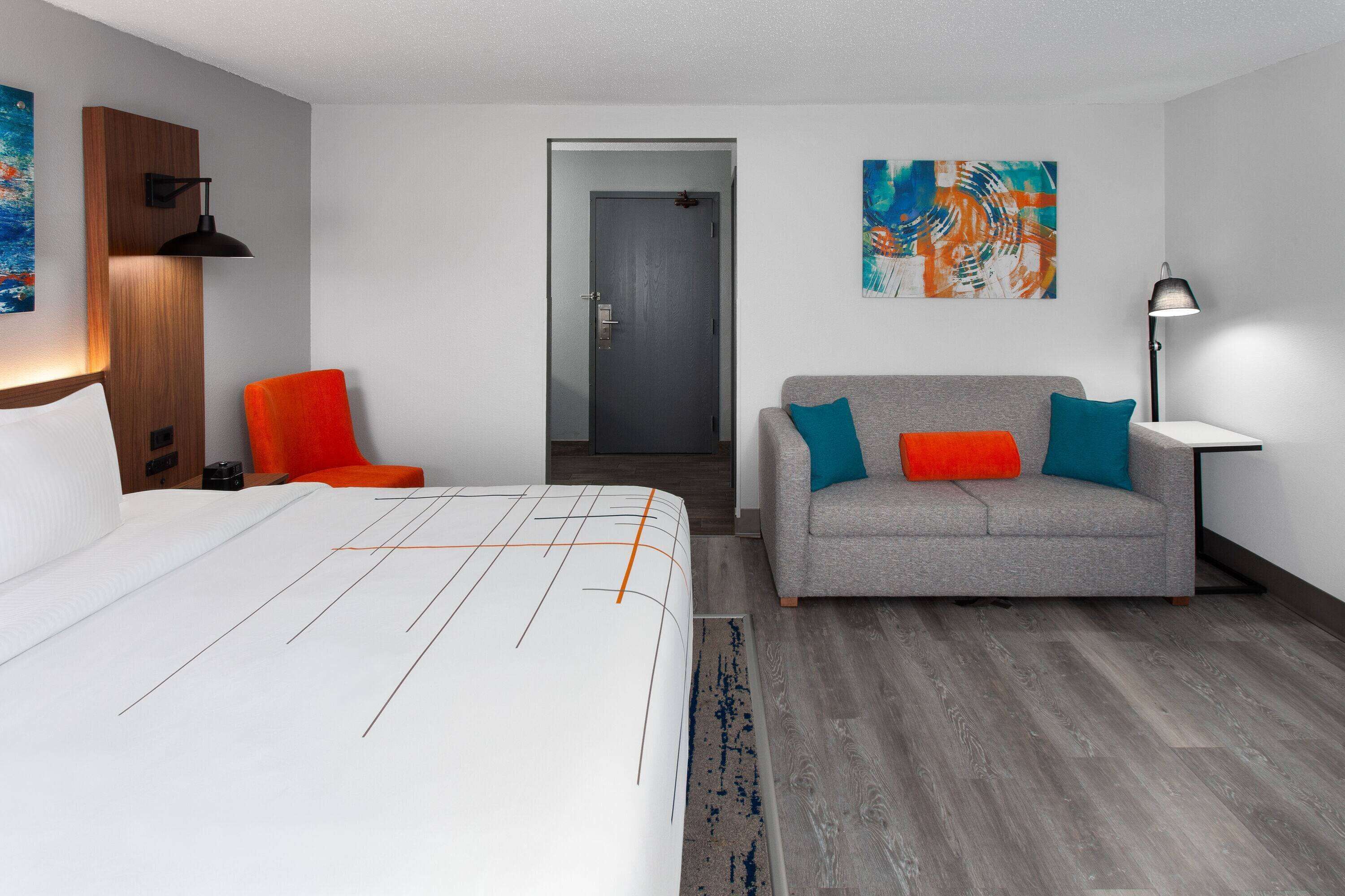 Une chambre d’hôtel avec un lit, un canapé gris agrémenté de coussins colorés, une chaise rouge, une tête de lit en bois et un tableau accroché au mur.
