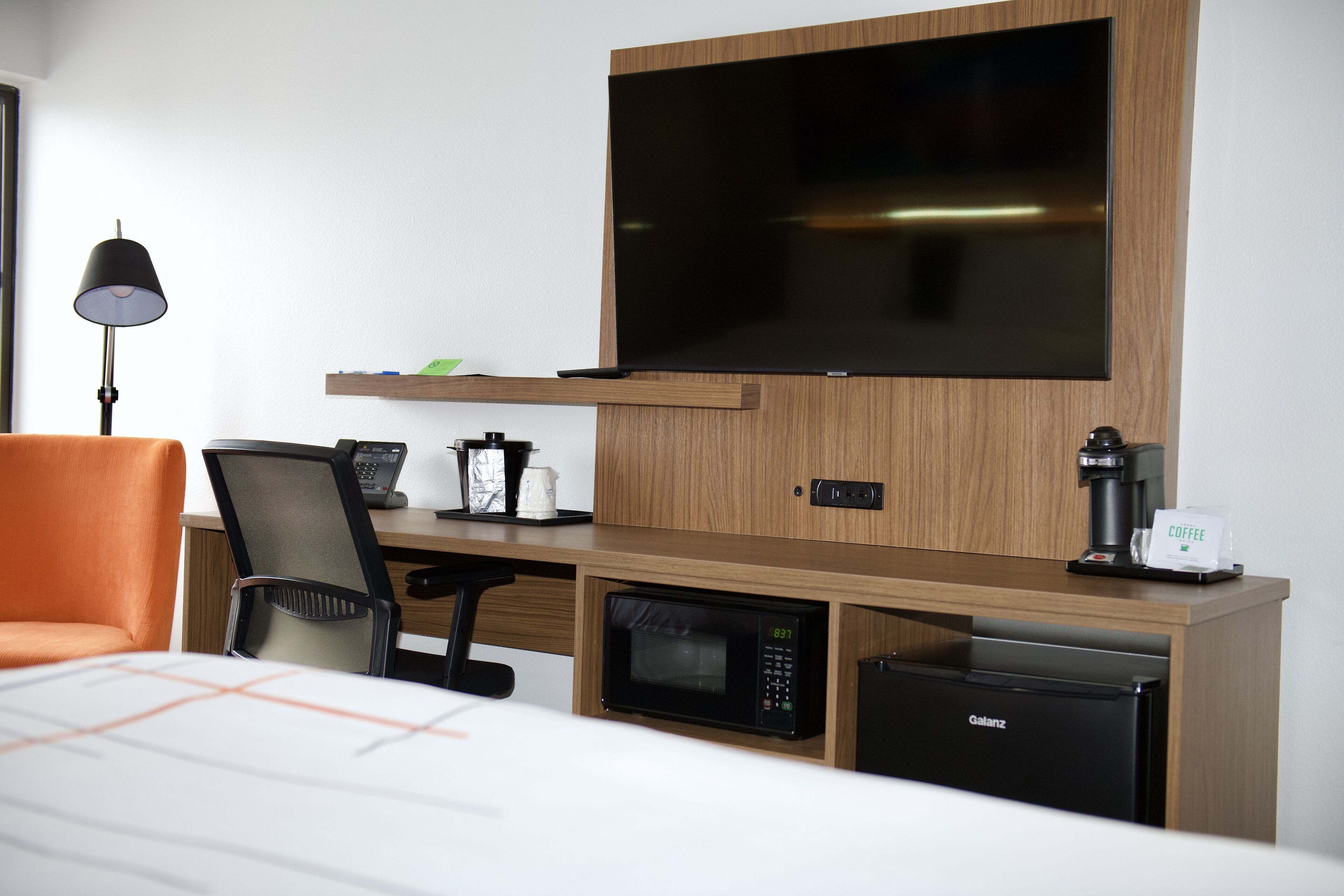 Ein Hotelzimmer mit einem Bett, einem Schreibtisch mit Computer, einer Mikrowelle, einer Kaffeemaschine, einem Fernseher und einer Lampe.