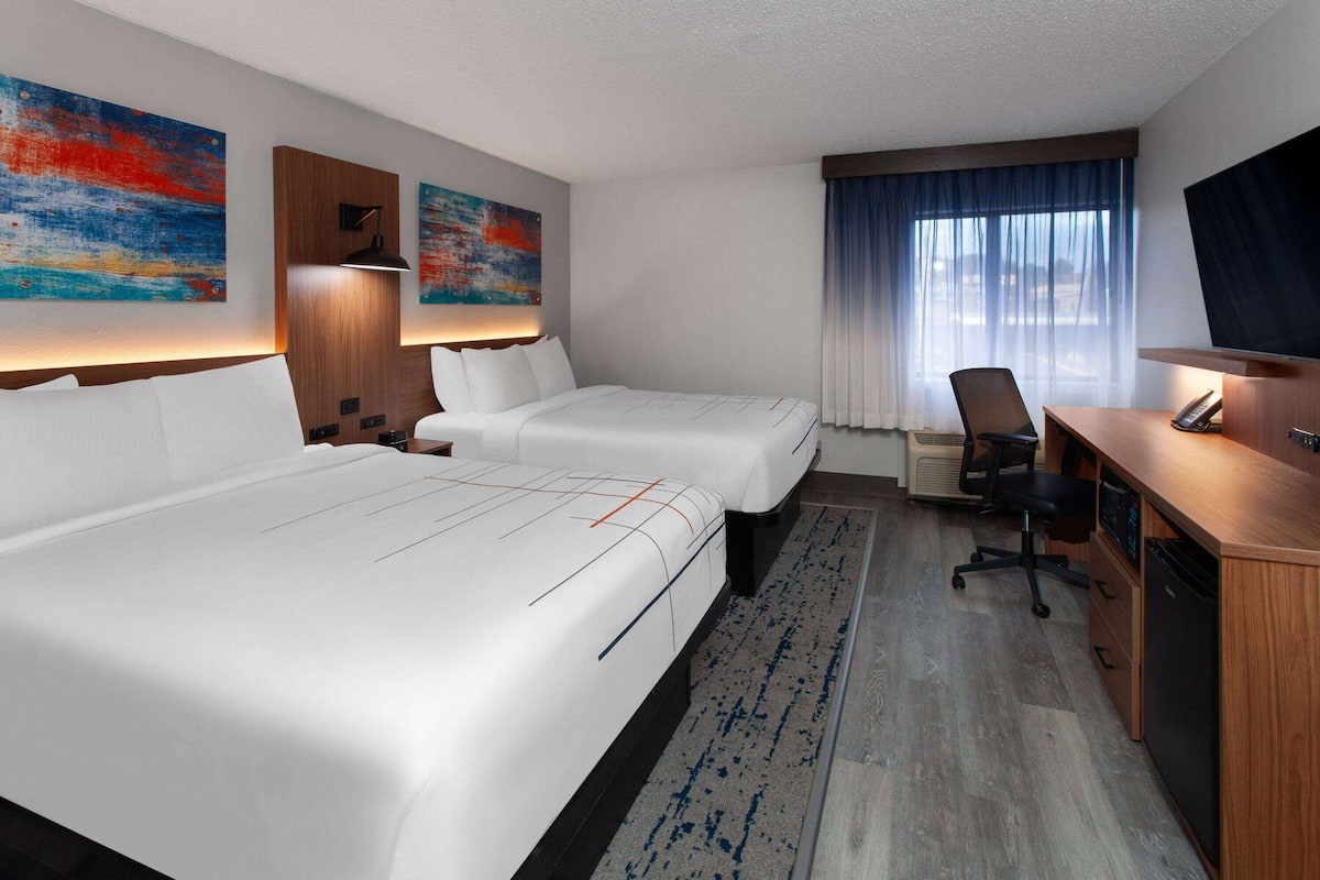 A hotel room with two beds, a desk, a chair, a TV, and artwork on the walls.