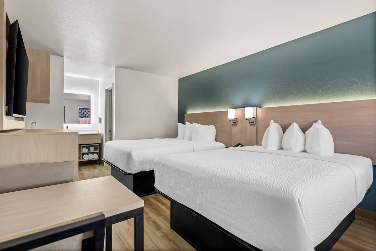 A hotel room with two beds, a desk, a TV, and a wall-mounted lamp.