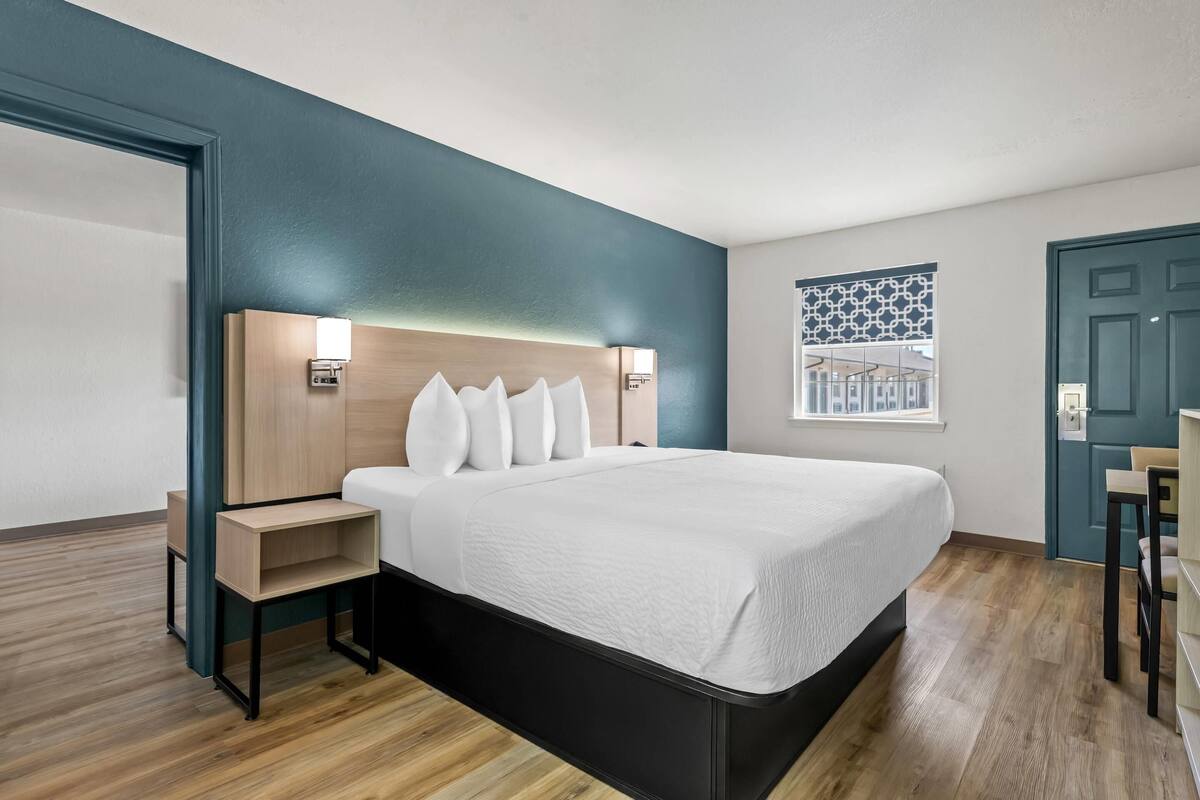 A modern hotel room with a large bed, built-in bedside tables, and a headboard with integrated lighting.