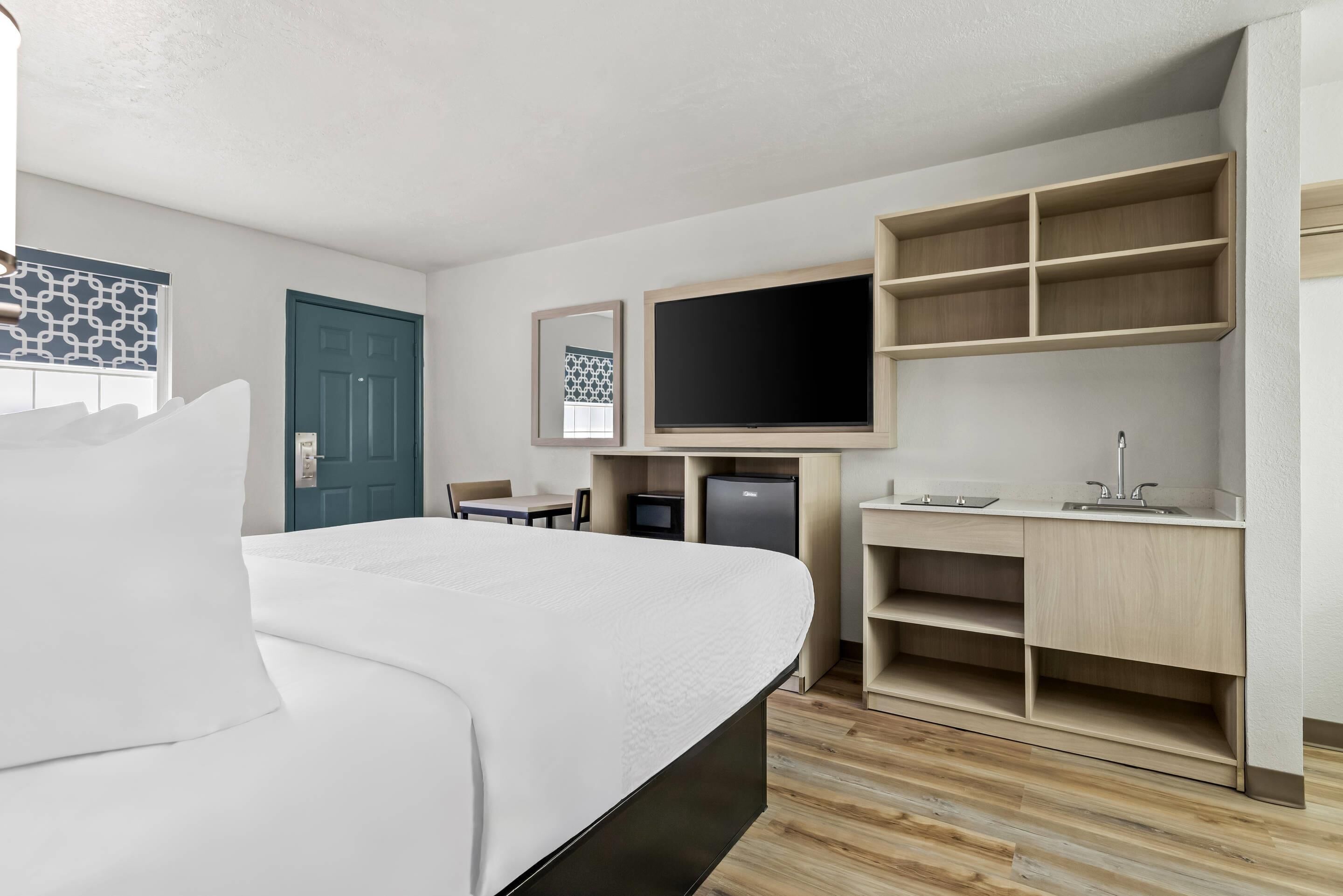 A modern hotel room with a large bed, a desk with a flat-screen TV, a small kitchenette, and a door.