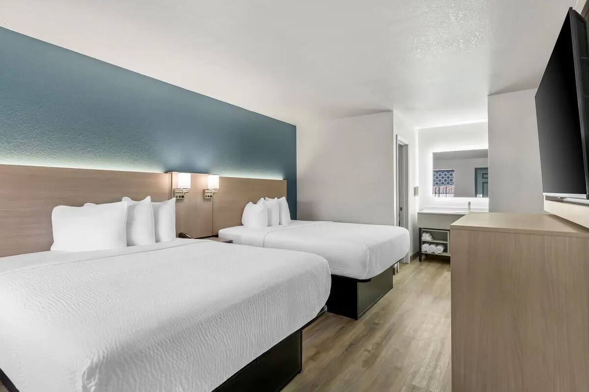 A hotel room with two beds, a desk, a TV, and a bathroom visible in the background.