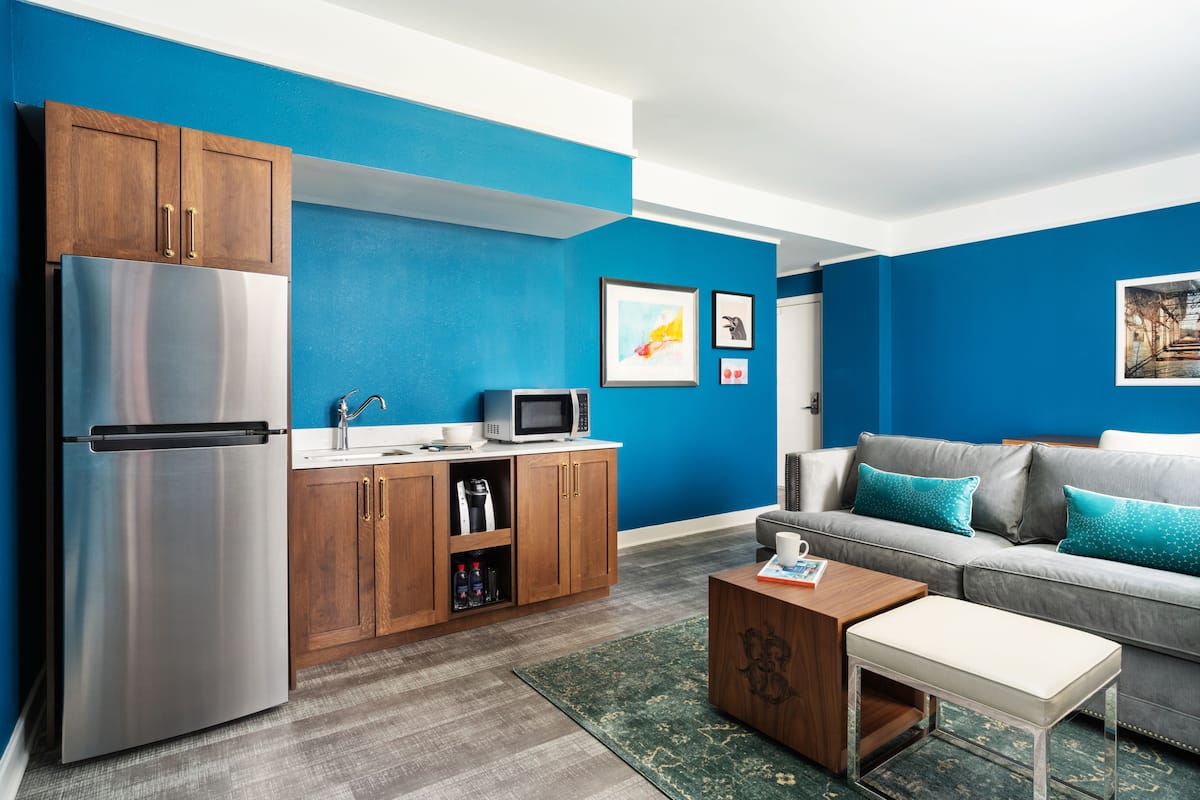 A modern kitchen with a stainless steel refrigerator, wooden cabinets, and a microwave. There is a grey sofa with teal cushions and a small wooden coffee table.