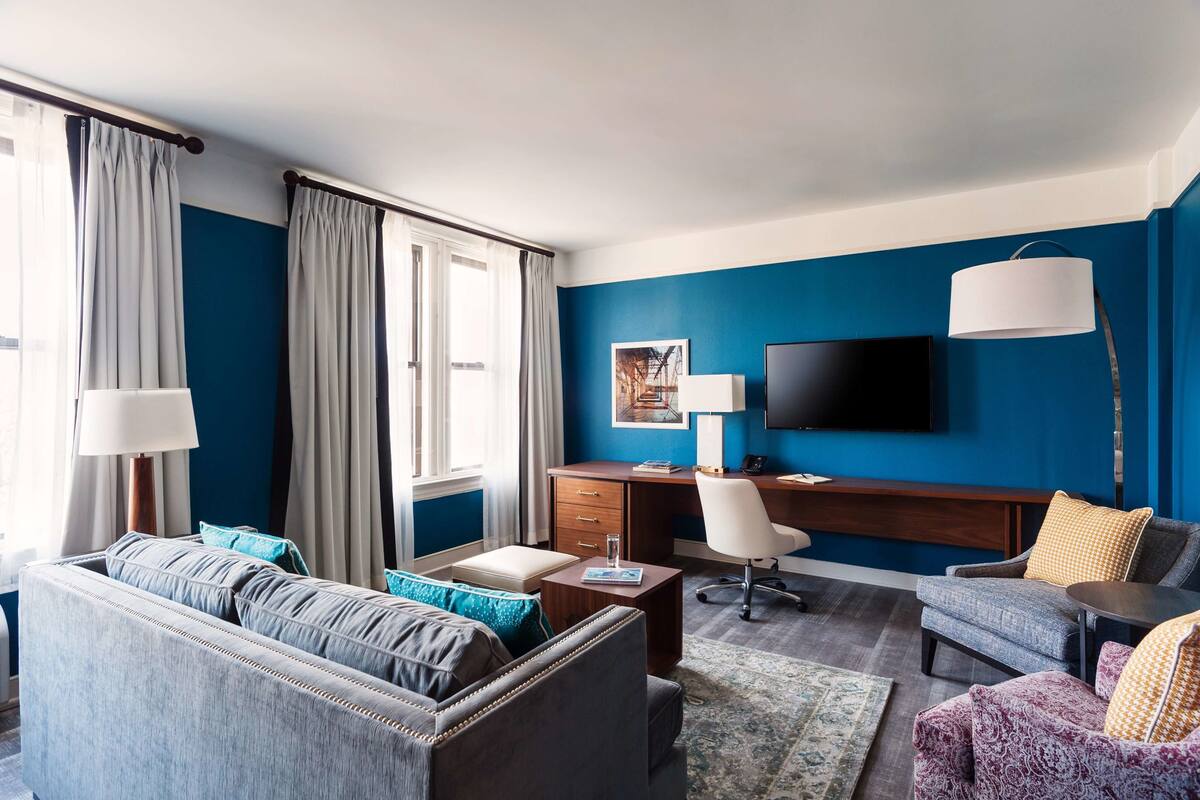A modern hotel room with a blue accent wall, a flat-screen TV, a desk with a chair, a sofa, and a coffee table.