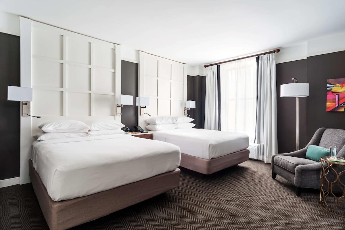 A hotel room with two beds, a chair, a table, and a large window with curtains.