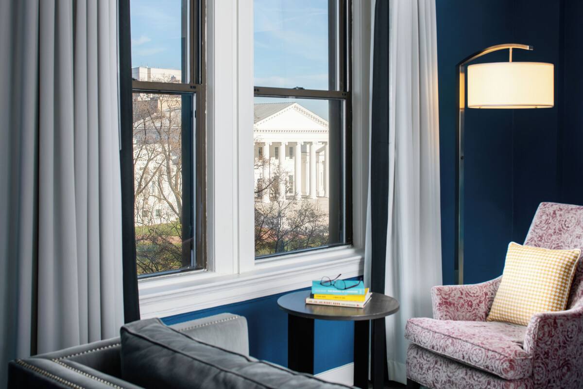 A room with a view of a neoclassical building through large windows, a sofa, a small table with books, and a patterned armchair.