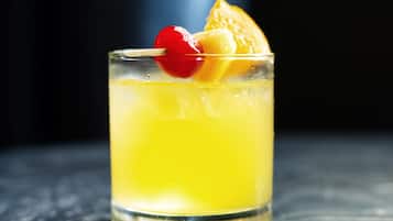 A glass of yellow beverage with an orange slice and a cherry on a stick.