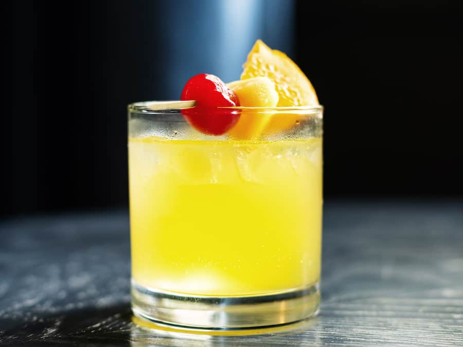 A glass of yellow beverage with an orange slice and a cherry on a stick.