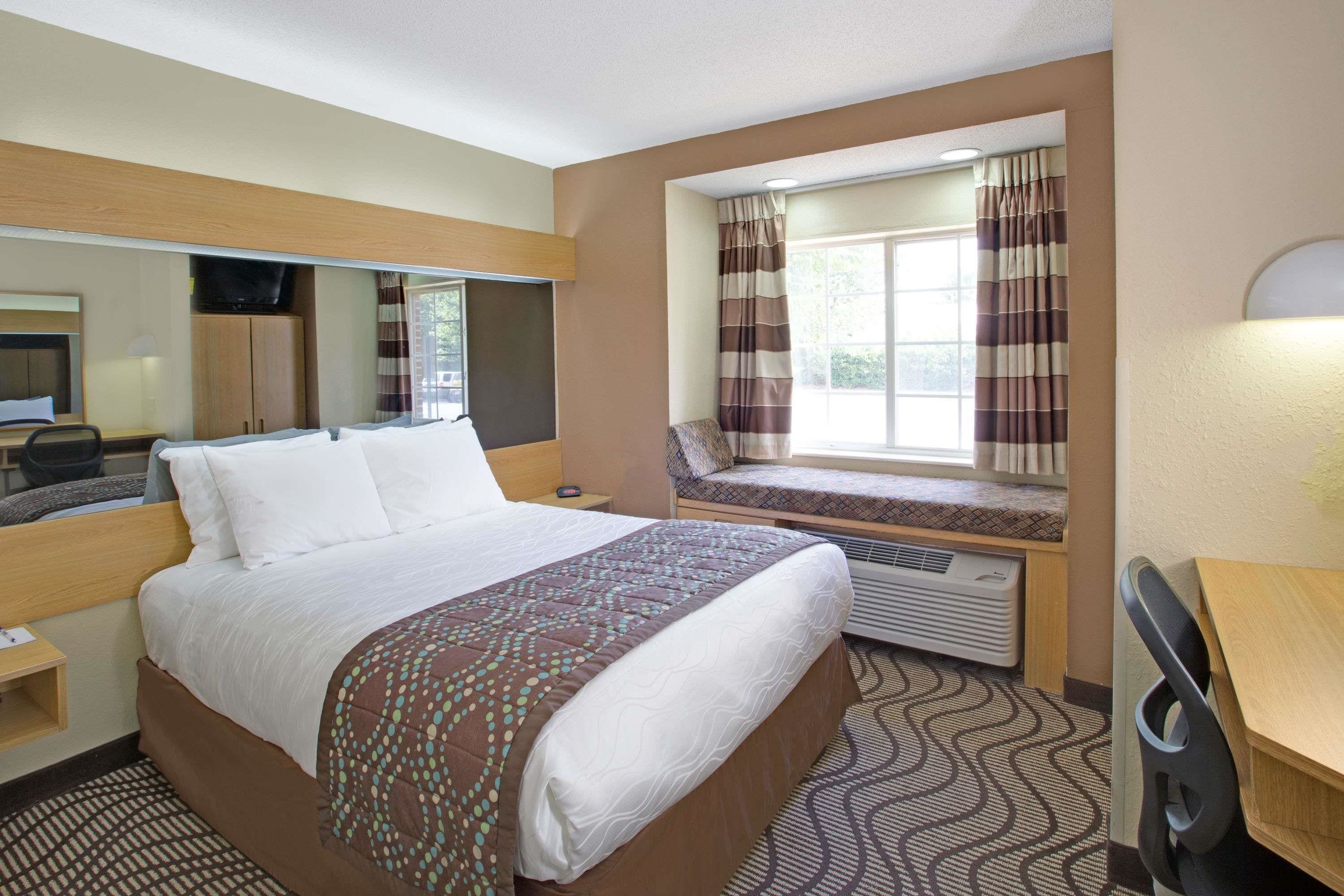 A hotel room with a large bed, a desk, a chair, a window with curtains, a television, and a heater.
