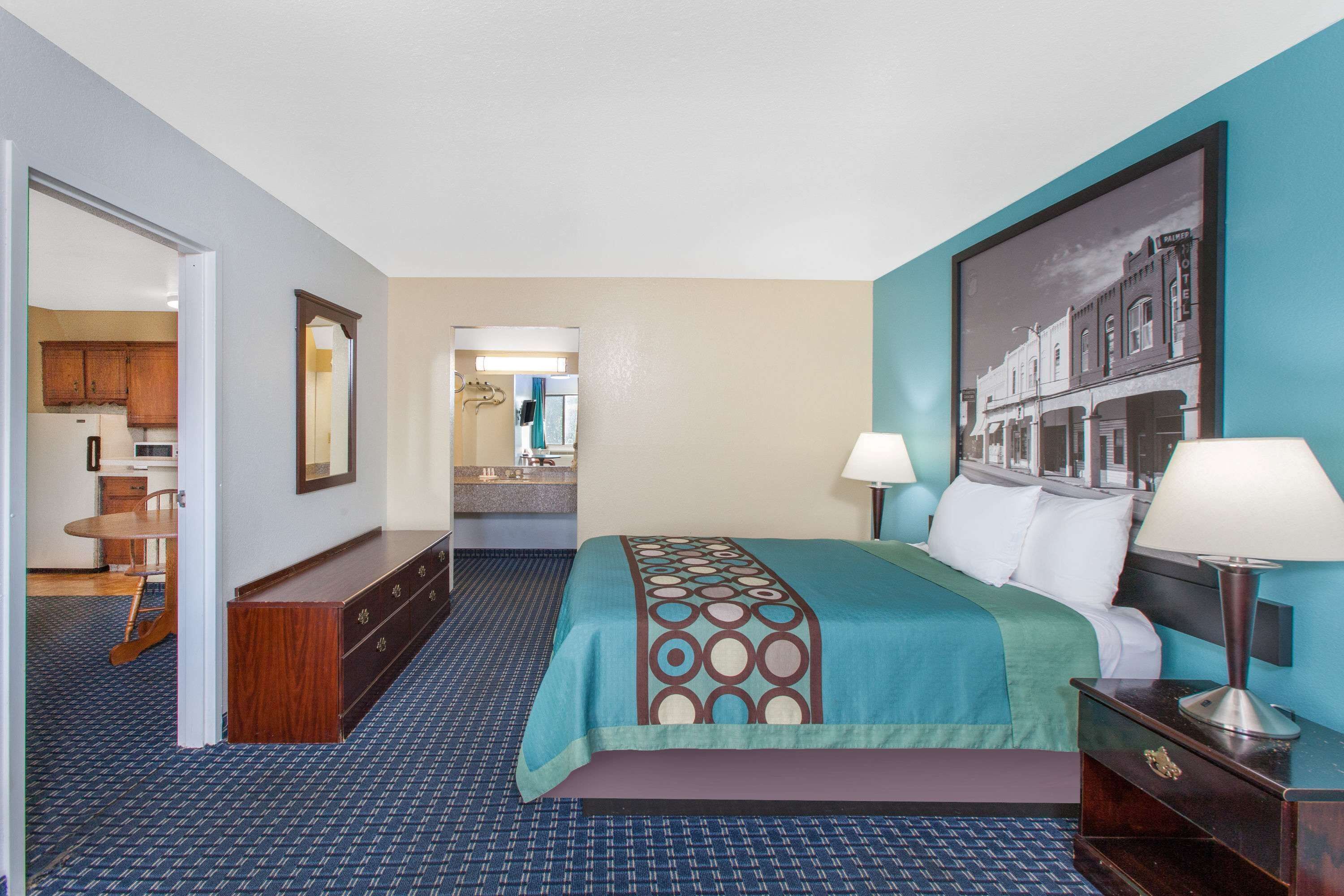 Habitación de hotel con cama, lámparas de noche, cómoda, espejo y vistas al pasillo y al baño.