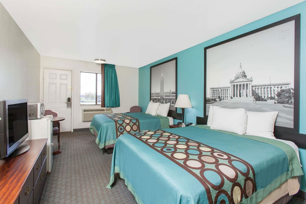 A hotel room with two beds, a desk with a TV, a window with curtains, and a large wall picture.