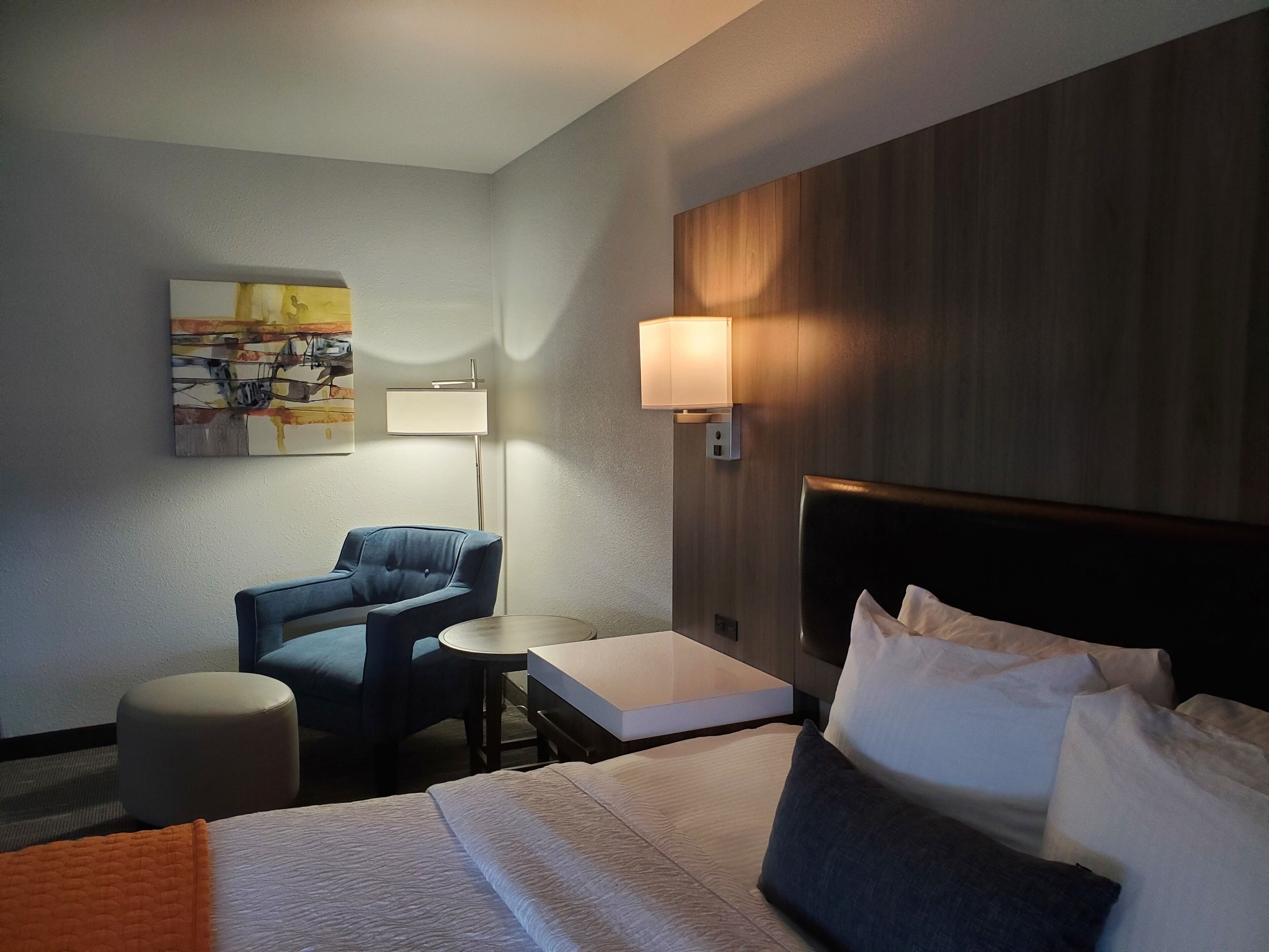 A hotel room with a bed, a desk, a chair, a lamp, and a painting on the wall.