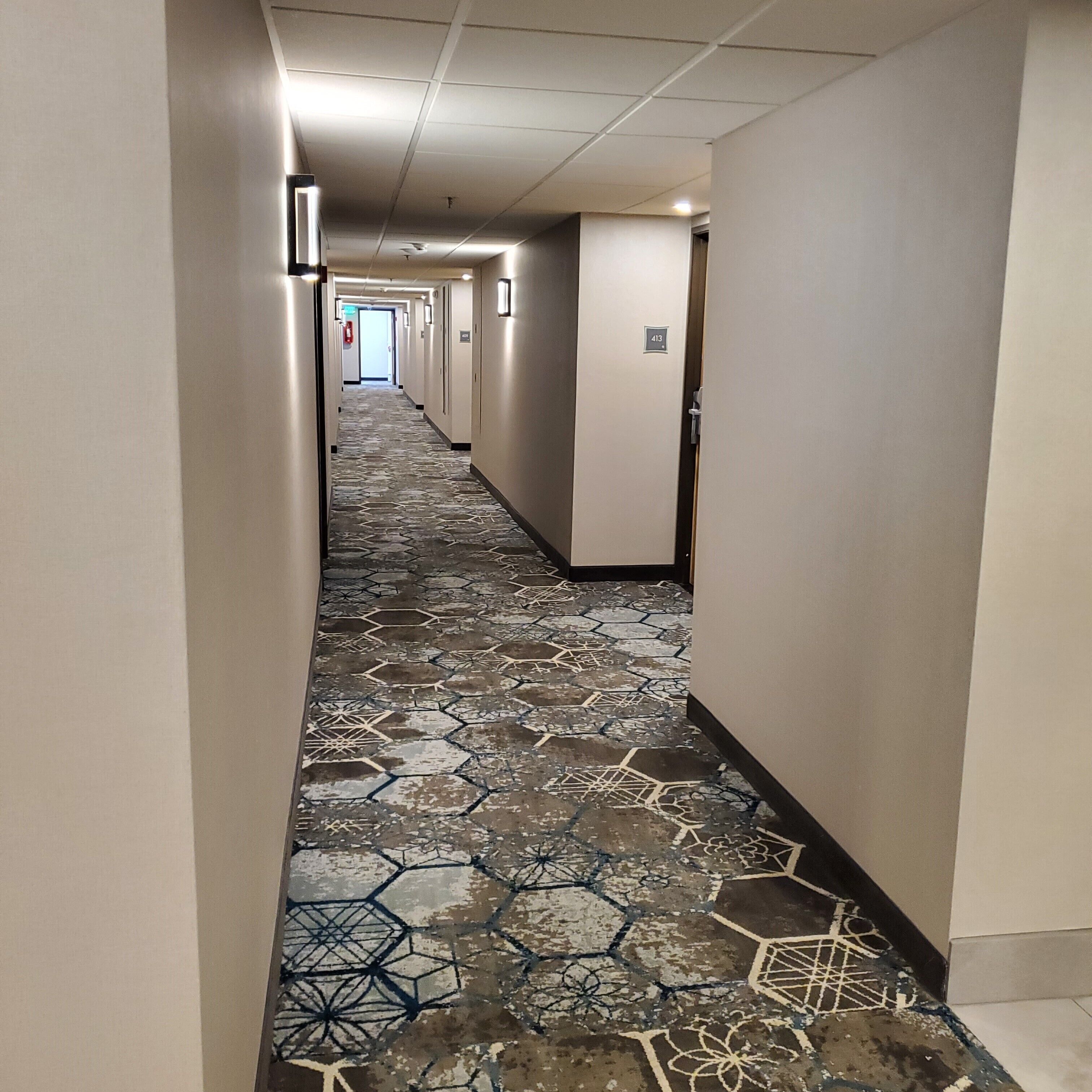 A hotel hallway with a patterned floor, doors on the right, and a fire extinguisher on the left.