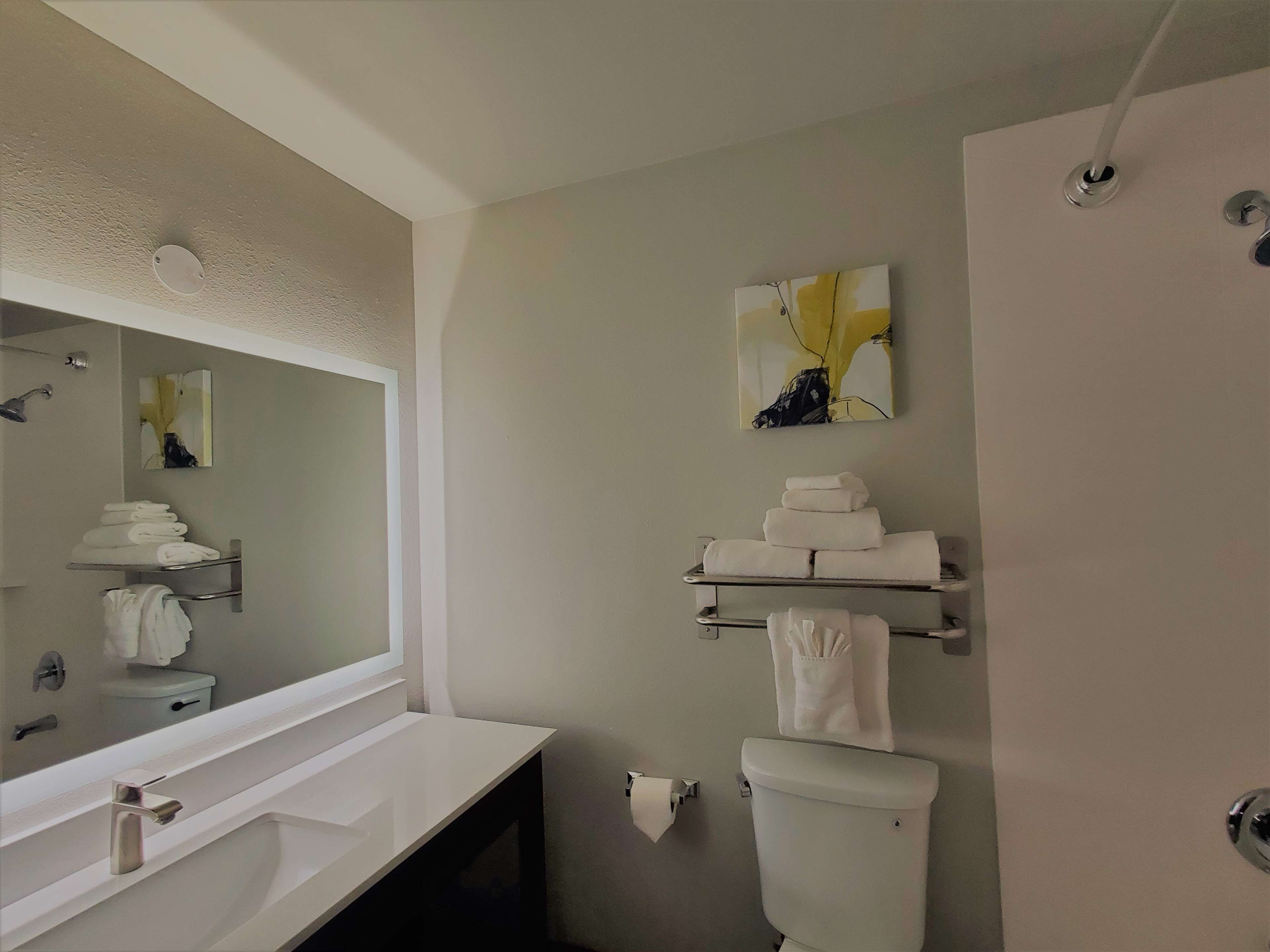 Et moderne badeværelse med en hvid vask, spejl og toilet. Der er håndklæder stablet på en hylde over toilettet.