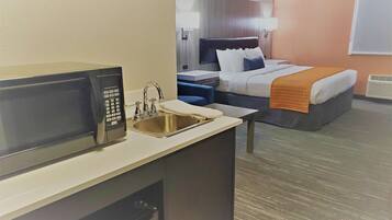 A hotel room with a kitchenette, a bed with a headboard, a bedside table, and a window.
