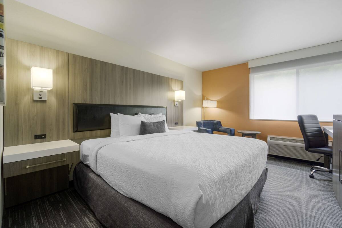 A hotel room with a large bed, bedside tables, a desk, a chair, and a window with blinds.