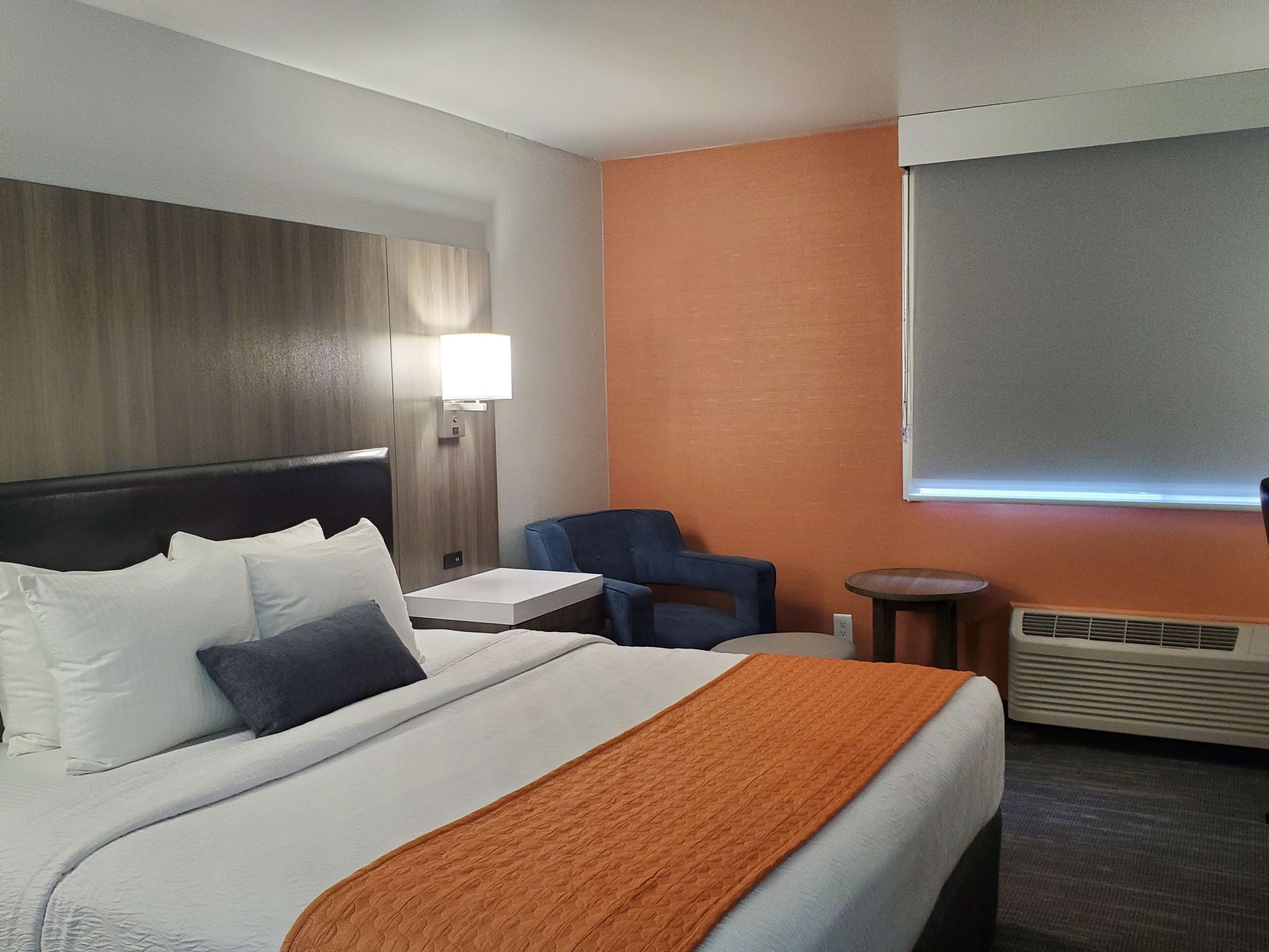 Une chambre d’hôtel avec un lit, une table de chevet, une chaise, une petite table et une fenêtre avec des stores.